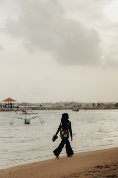A woman walks along a tranquil beach in Bali, Indonesia, during dusk, with soft waves and a cloudy sky.