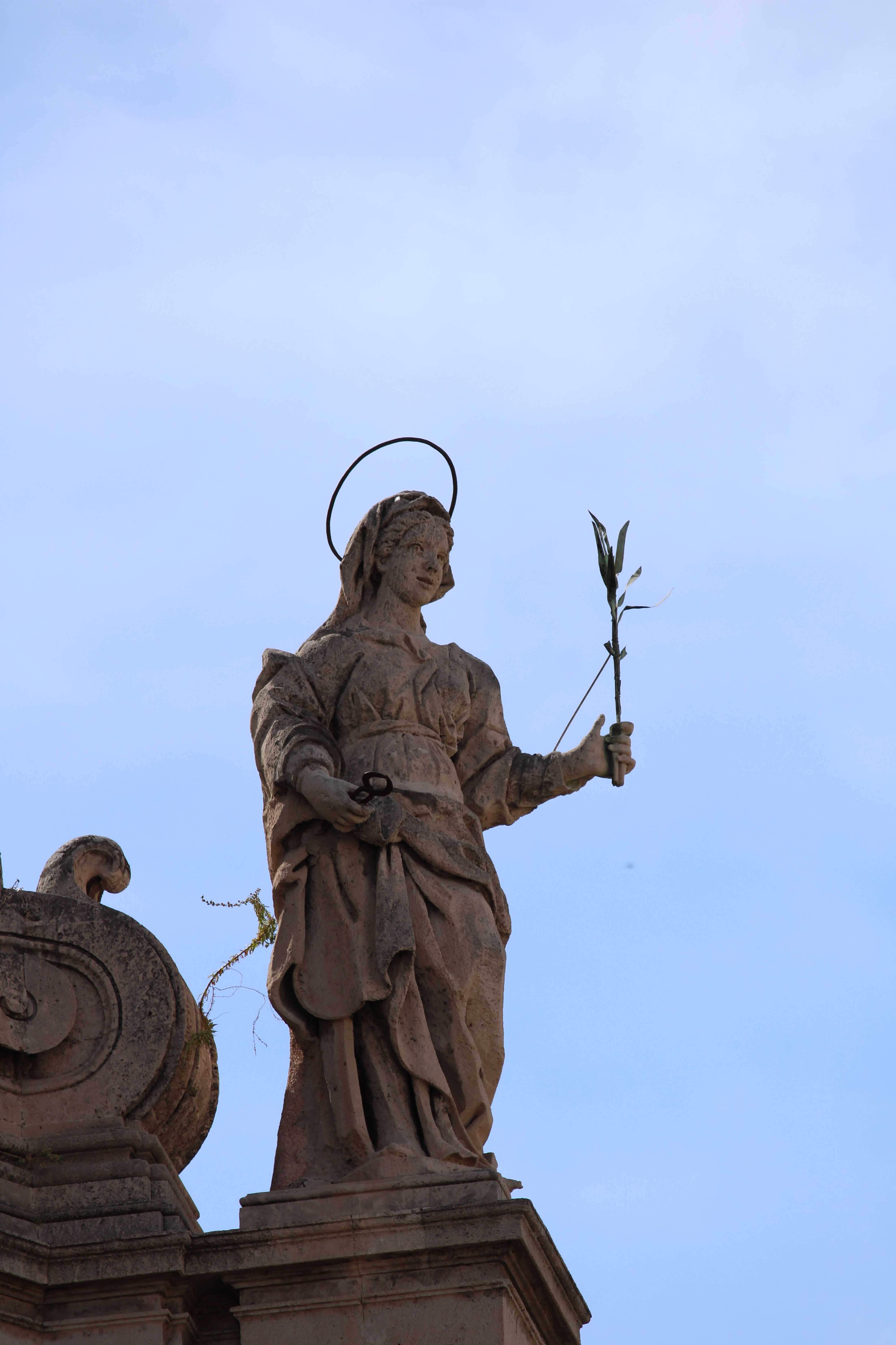 Elegant stone statue with halo holding a branch on a classic architectural pedestal against the sky.