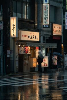 Cozy, rainy Tokyo street with glowing signs, umbrellas, and reflections on wet pavement.