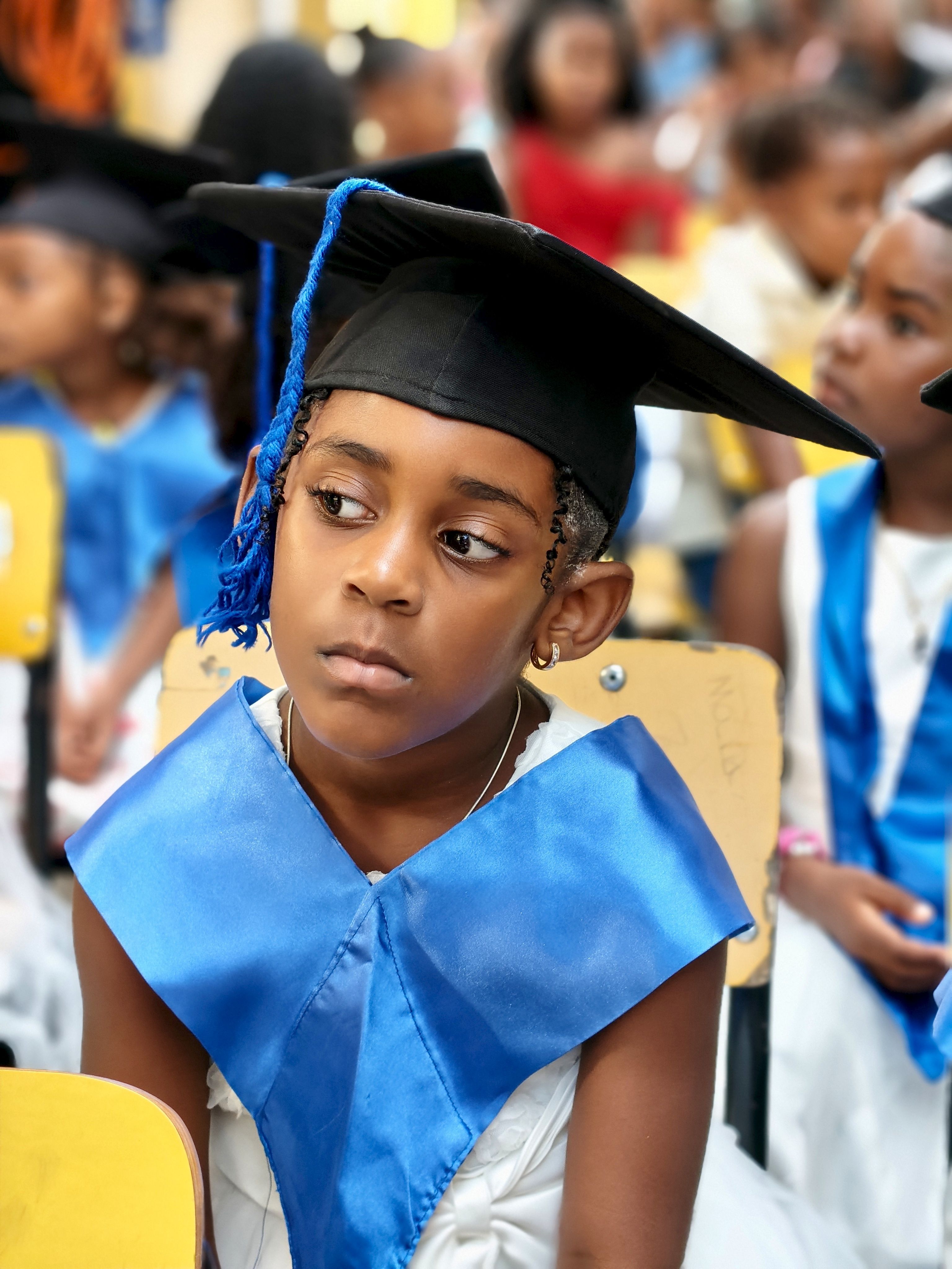 A young girl in a blue graduation robe and cap attending a school graduation ceremony.