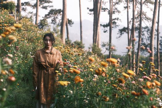 Woman in trench coat stands among colorful flowers in wooded meadow.