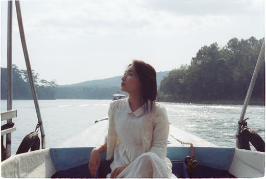 Woman in a white dress sits on a boat in a tranquil lake surrounded by trees.