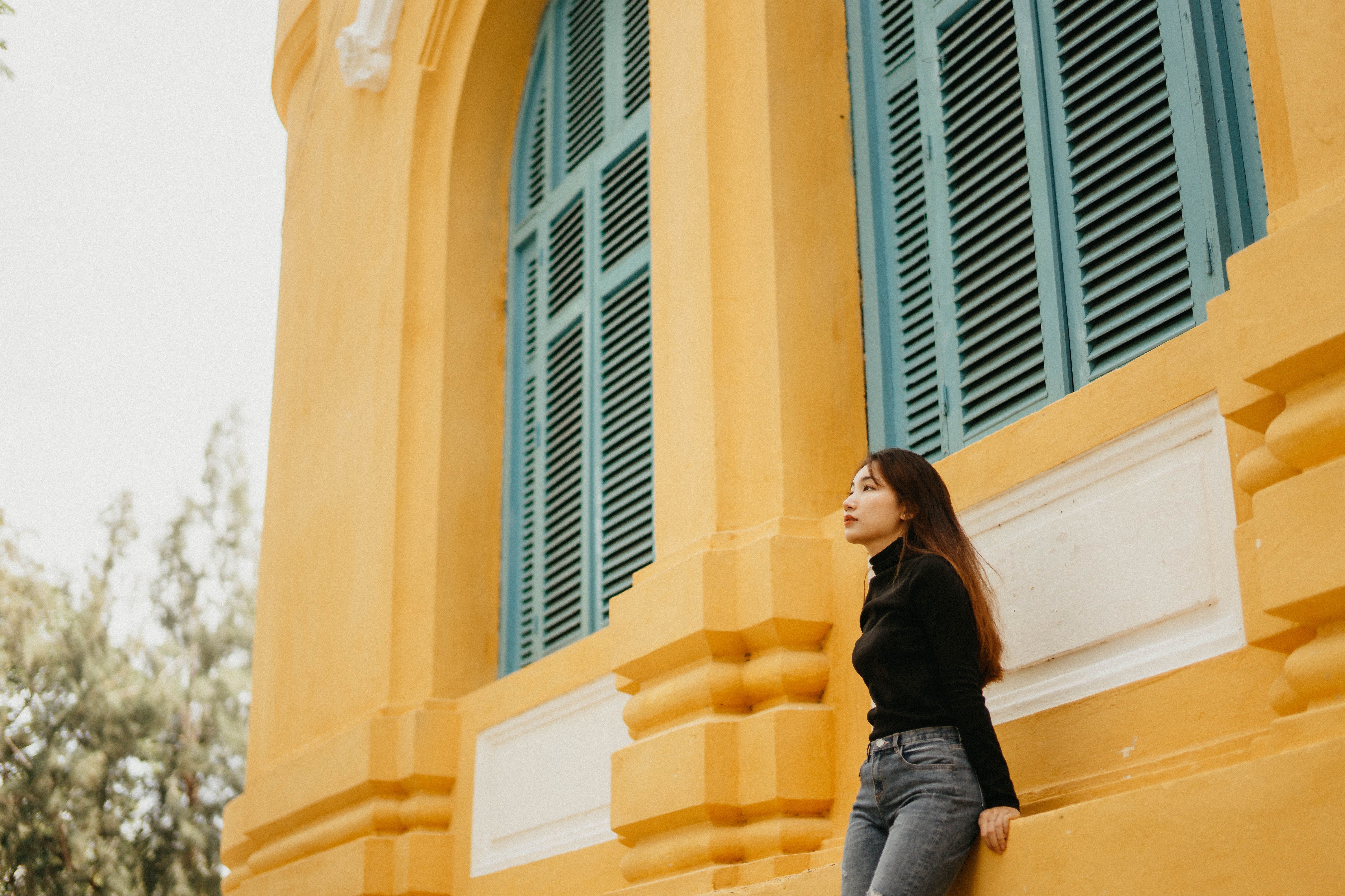 A young woman in casual attire leans against a colorful yellow wall with teal windows outdoors.