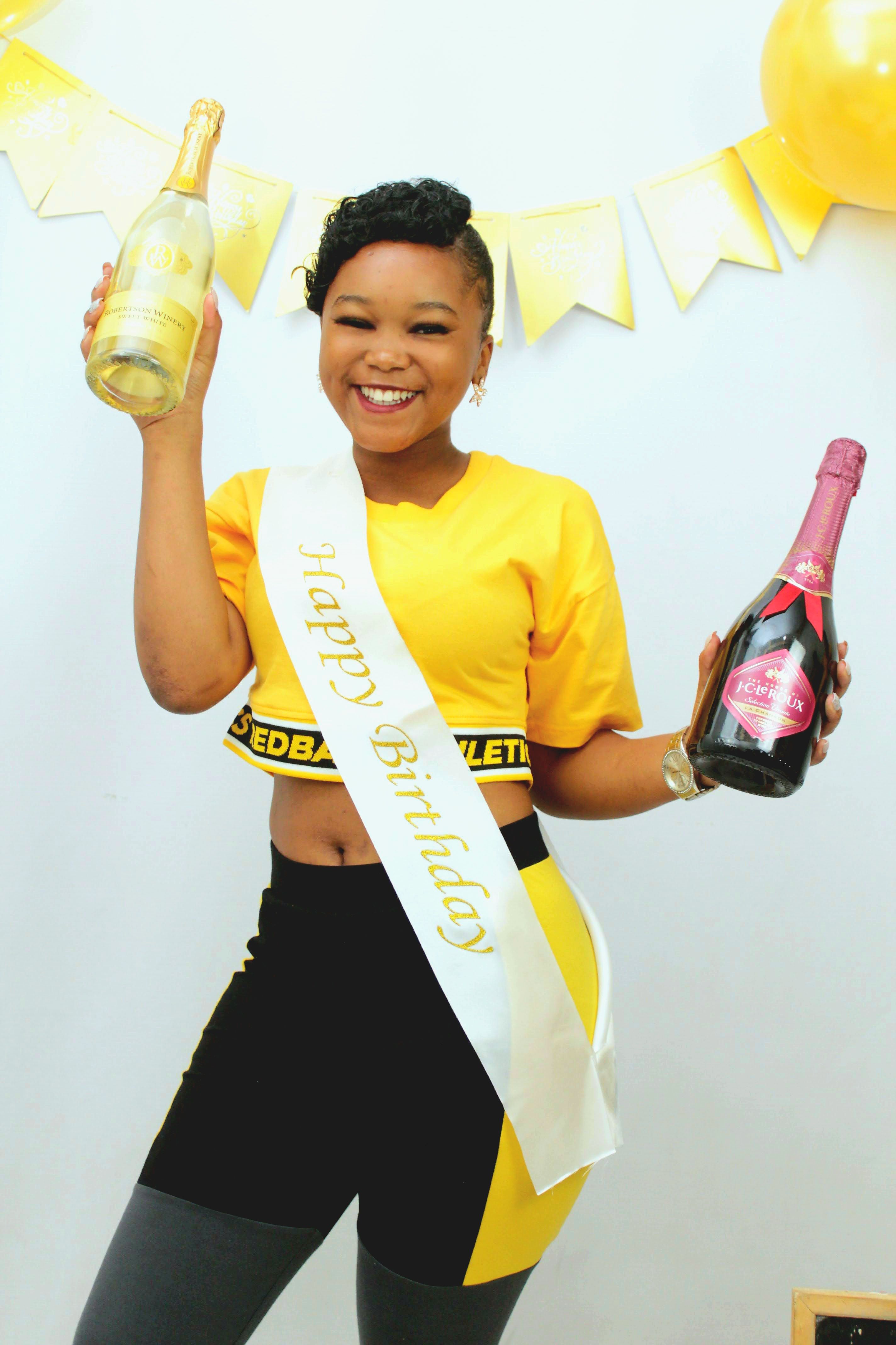 Smiling young woman with champagne bottles and a birthday sash celebrating indoors.