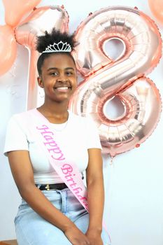 Smiling young woman celebrating her 18th birthday with balloons and a birthday sash.