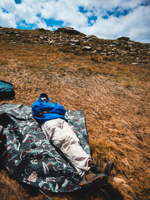 Free A person in outdoor gear relaxing on a patterned blanket on a grassy mountain slope under a cloudy sky. Stock Photo