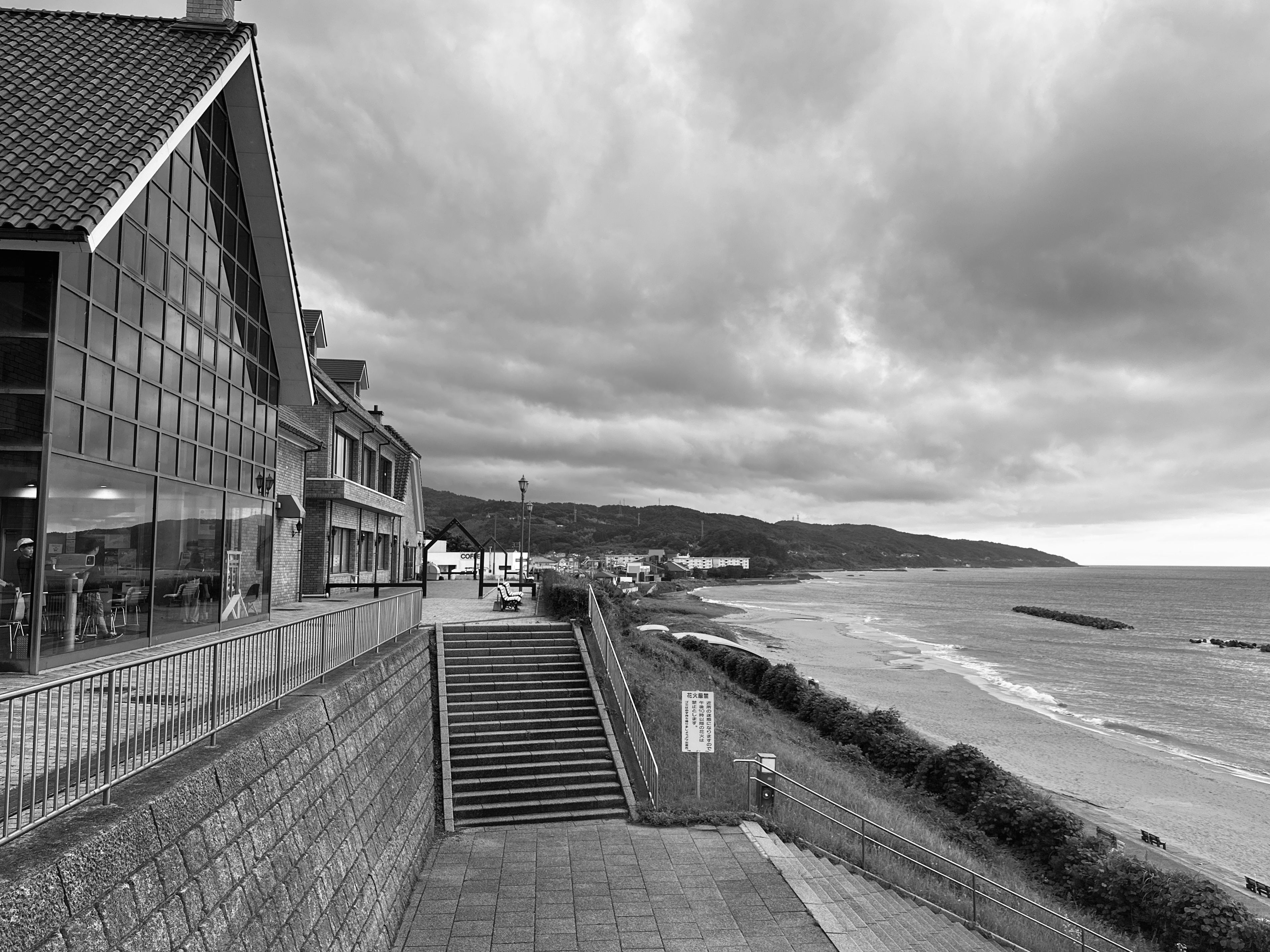 近代的な建物とドラマチックな曇り空が特徴的な海岸風景を写した白黒写真。