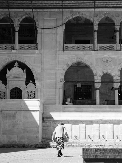 Free A woman walks in front of a historic stone building with arches, creating a classic monochrome scene. Stock Photo