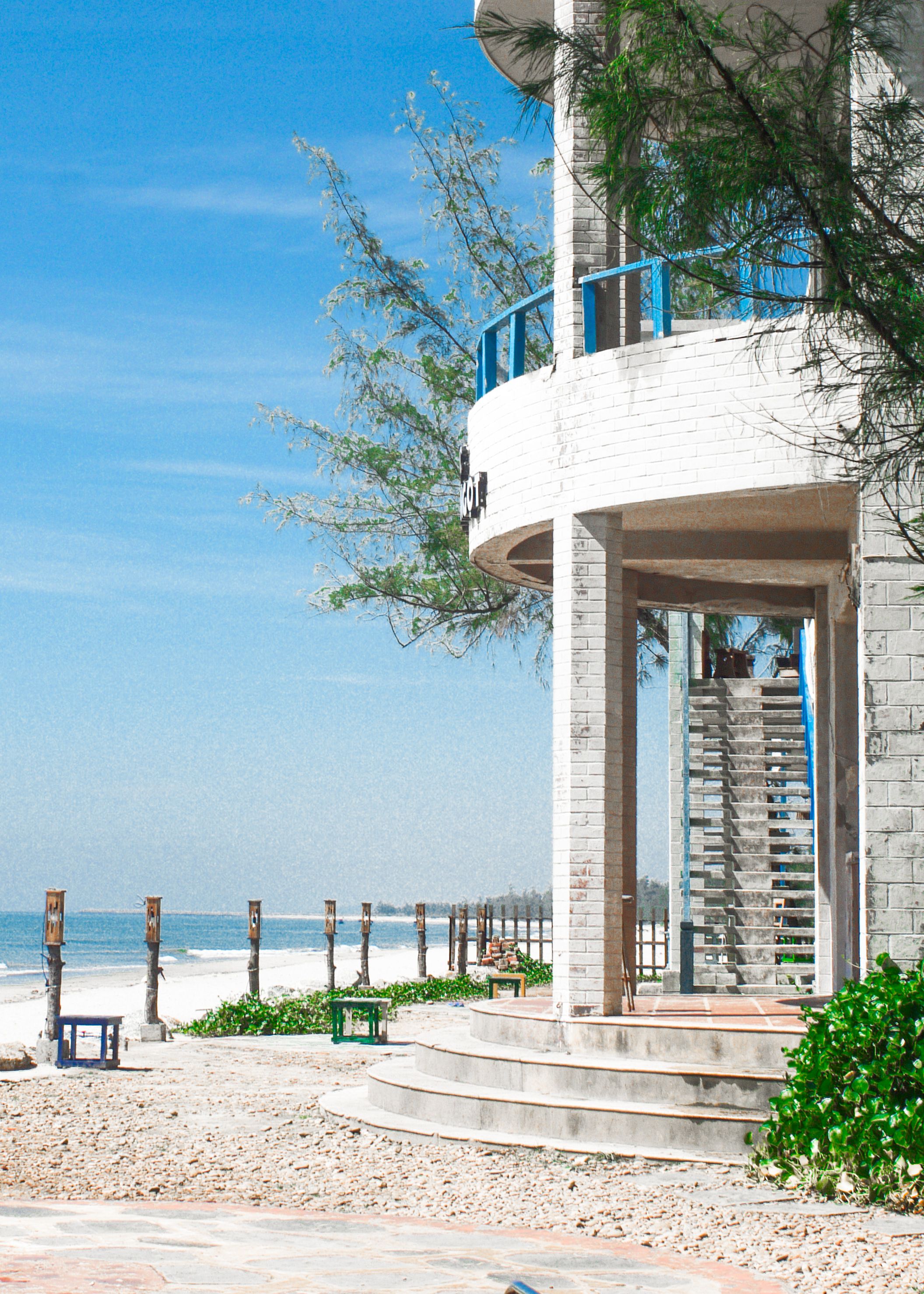 Charming coastal building with stairs and railings overlooking a sandy beach under a bright blue sky.