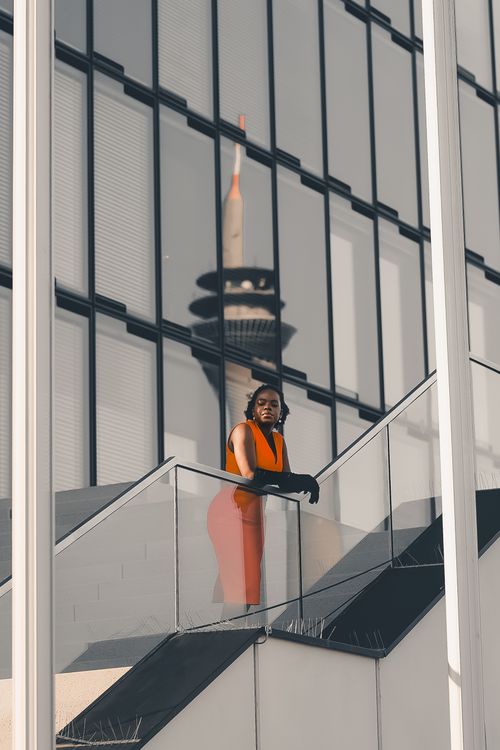 Free A fashionable woman in vibrant attire poses on a modern staircase with a tower reflection. Stock Photo