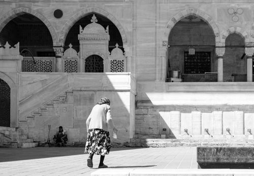 Free A monochrome photo of an old woman walking in front of historic stone arches. Stock Photo