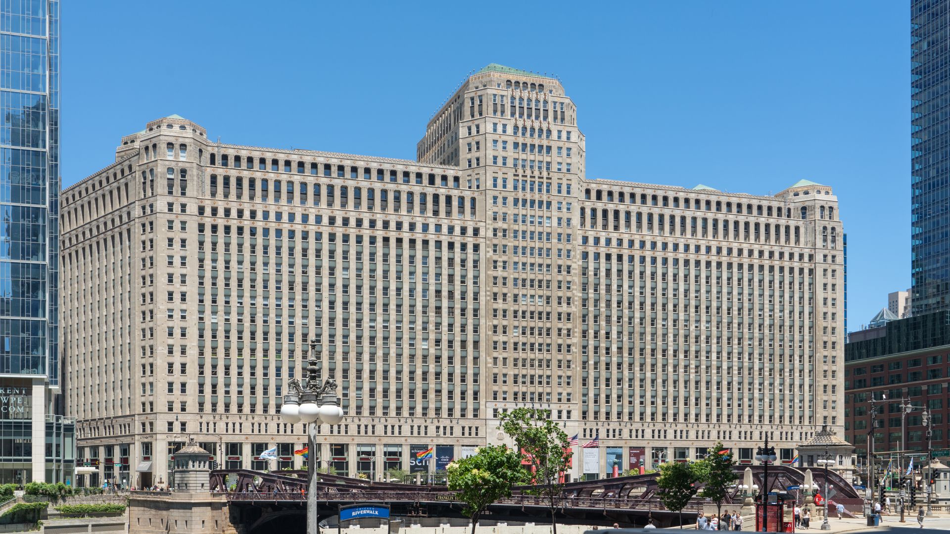 The Merchandise Mart building in Chicago, a historic beige Art Deco structure along the Chicago River with modern skyscrapers nearby under clear blue sky