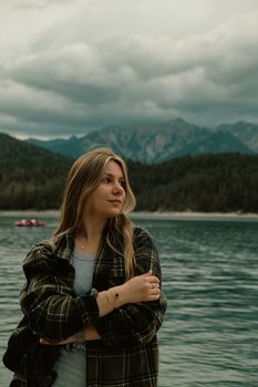 A serene portrait of a young woman standing by a picturesque mountain lake with lush forests.