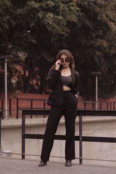 Woman in stylish black suit poses confidently outdoors with sunglasses in an urban environment.