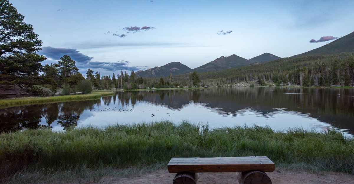 Peaceful sunrise at a mountain lake with a bench overlooking calm reflective waters and scenic mountains.