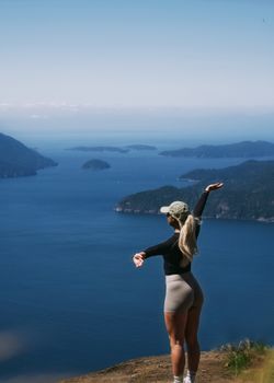 A woman celebrates at a mountain peak, arms wide open overlooking the ocean and islands.