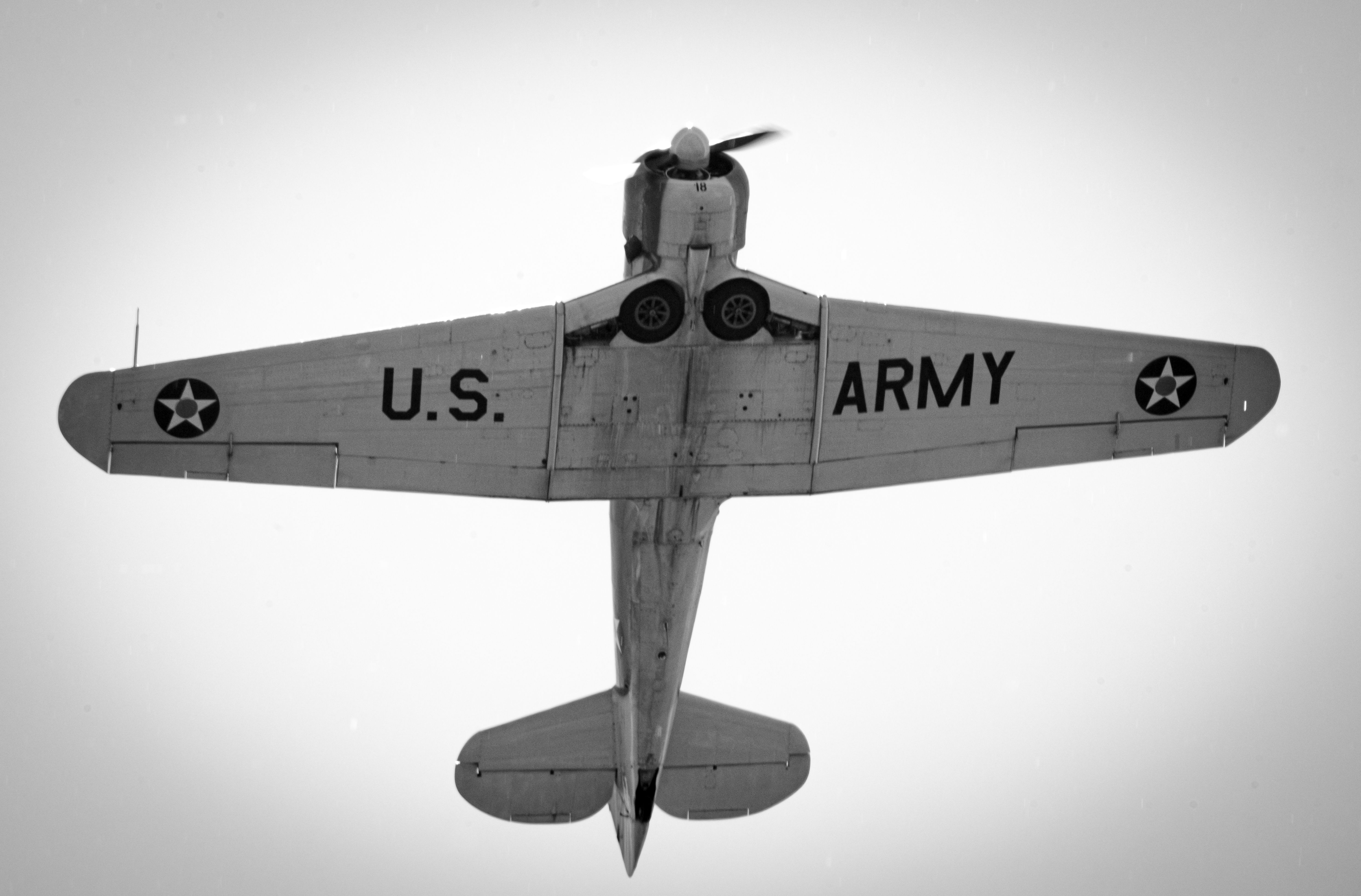 Black and white image capturing the underside of a vintage U.S. Army aircraft in flight.