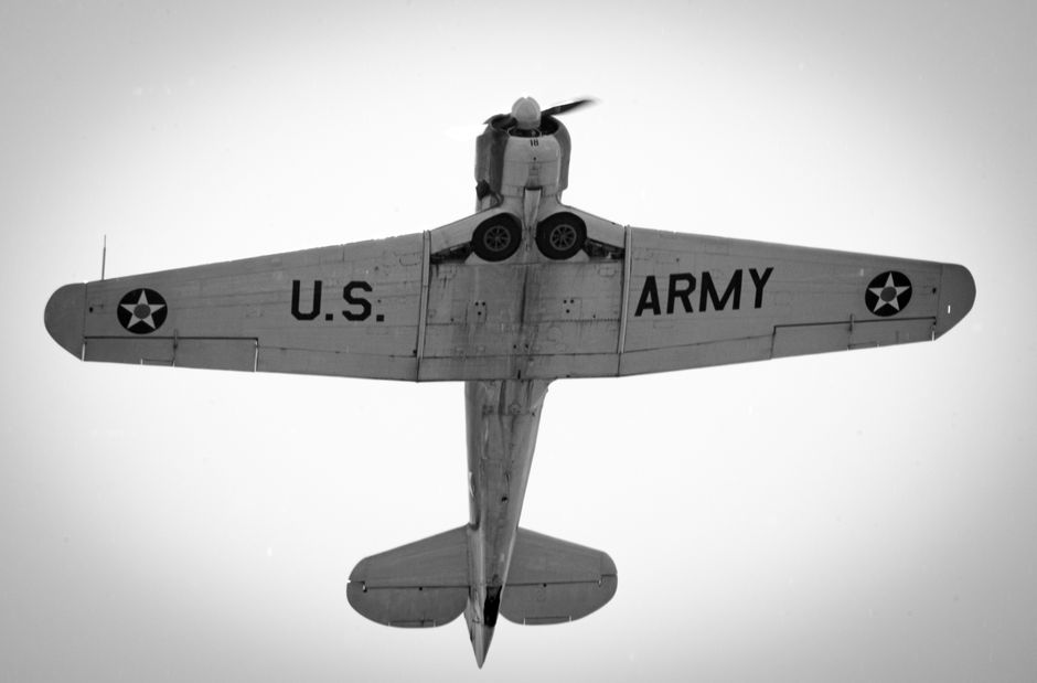 Black and white image capturing the underside of a vintage U.S. Army aircraft in flight.