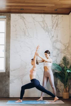 A yoga instructor helps a woman perfect her Warrior I Pose indoors.