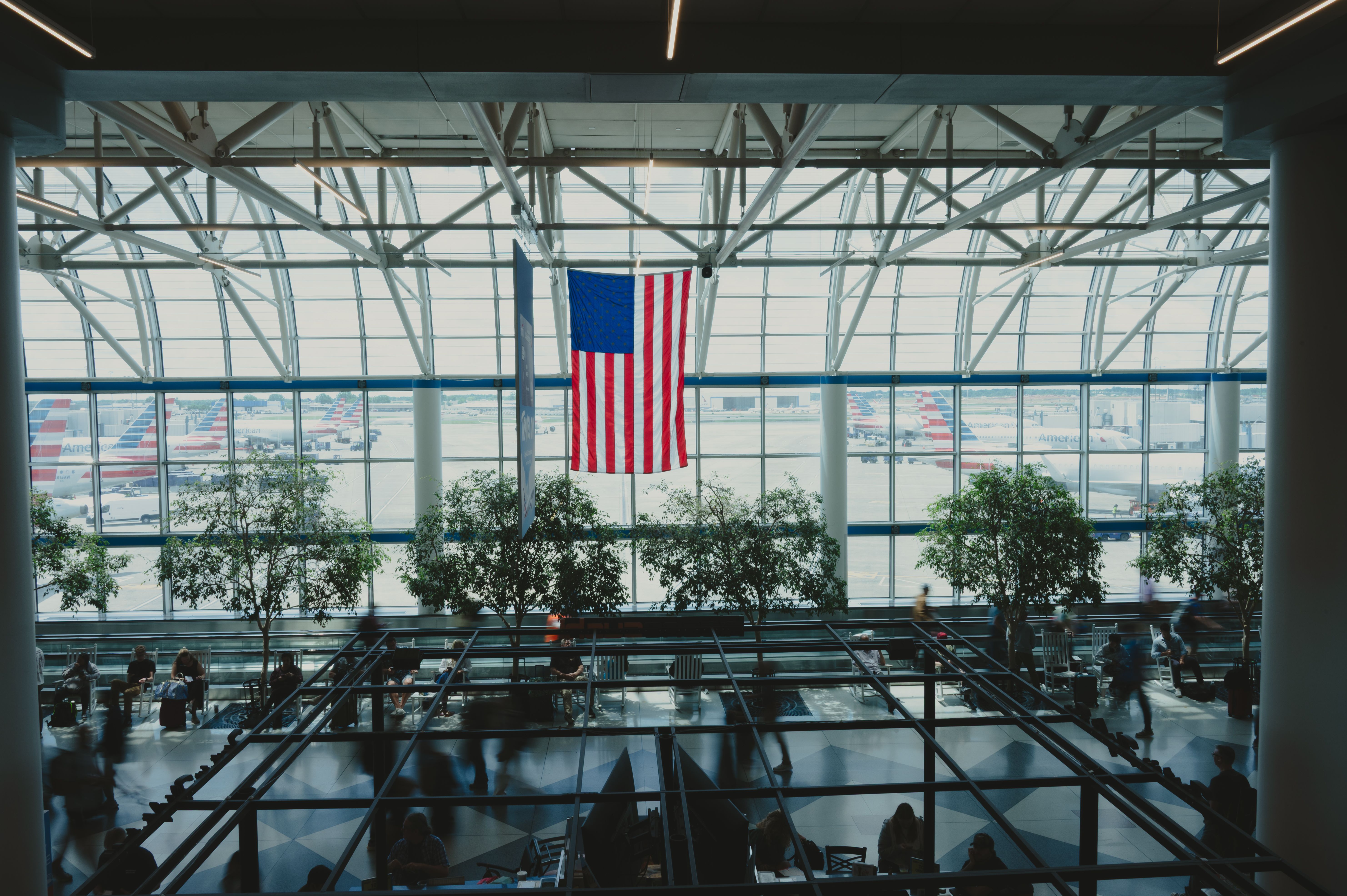 Interior view of an airport terminal's glass facade with an American flag, trees, and travelers.