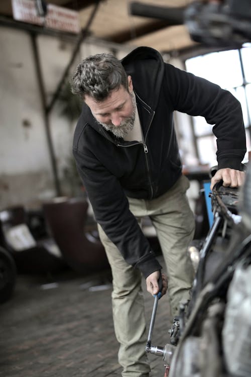 Mature male worker in casual clothes tighten nut with ratchet and repairing mean of transport while working in workshop