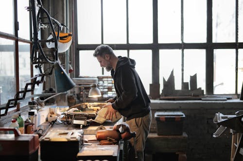 Side view of male workshop owner standing near messy workbench and fixing metal details while using instruments during work