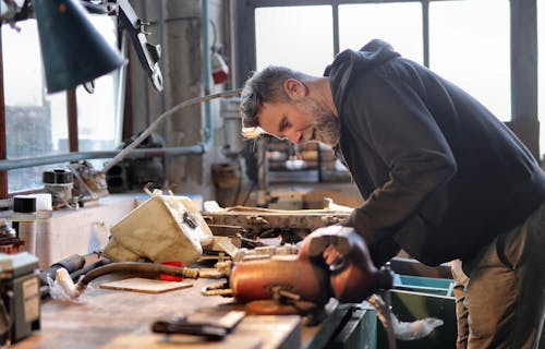 Free Side view of brutal male worker in casual wear standing at workbench and fixing metal details while using tools and working in workshop Stock Photo
