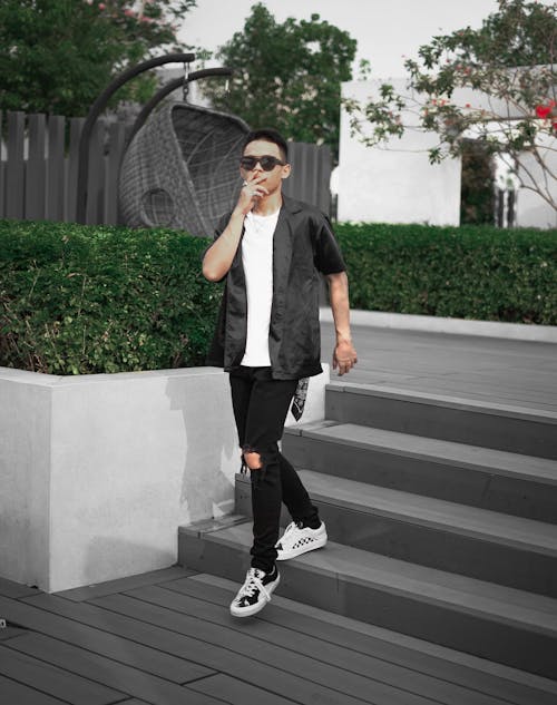 Full body of fit anonymous man in stylish wear and different sneakers touching face while walking down clean staircase near hedge