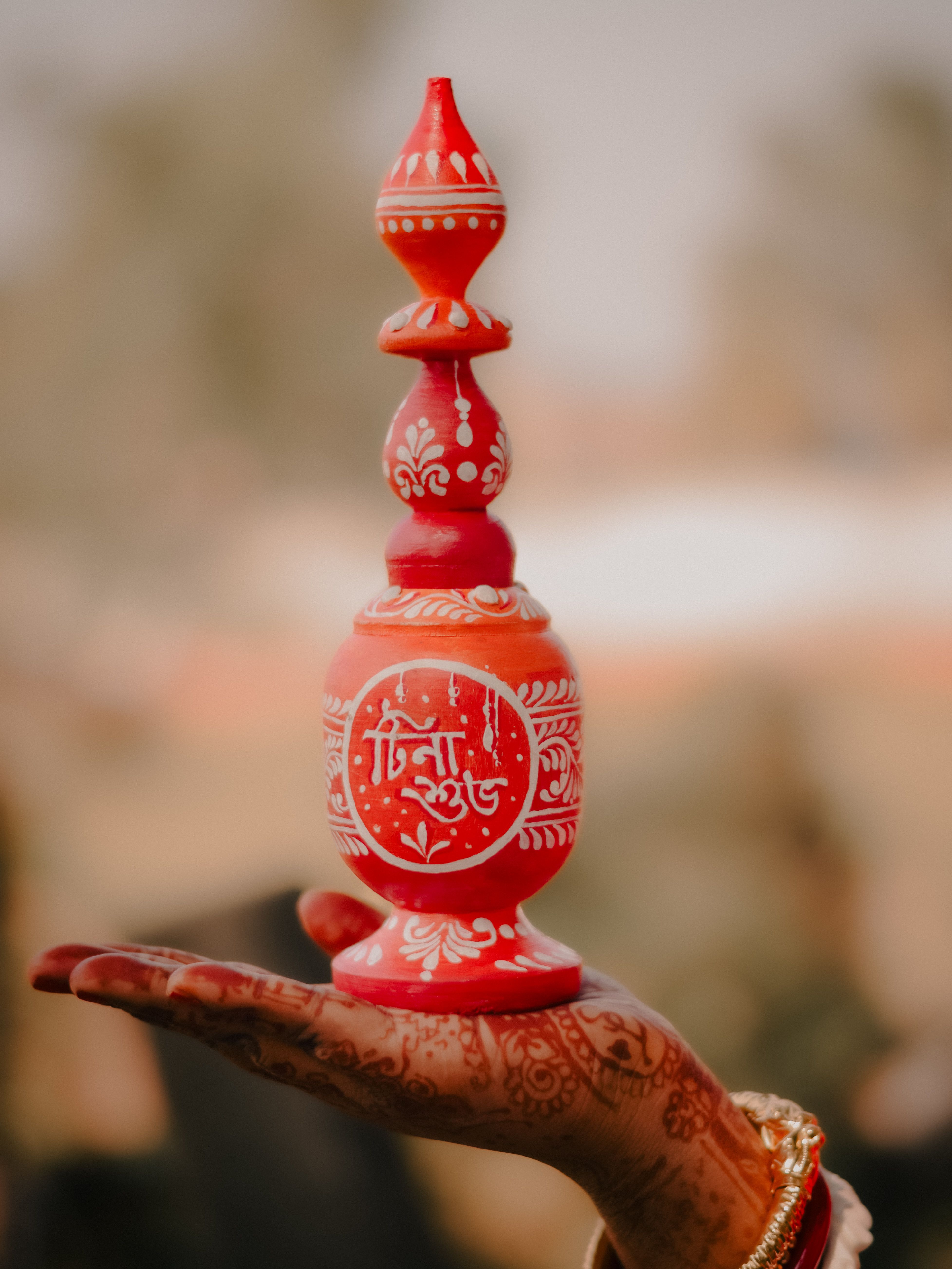 Elegant red gamcha pot delicately perched on a mehndi-decorated hand, symbolizing Bengali cultural heritage.