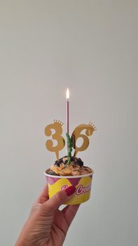 Hand holding ice cream cup with candle celebrating 36th birthday indoors.