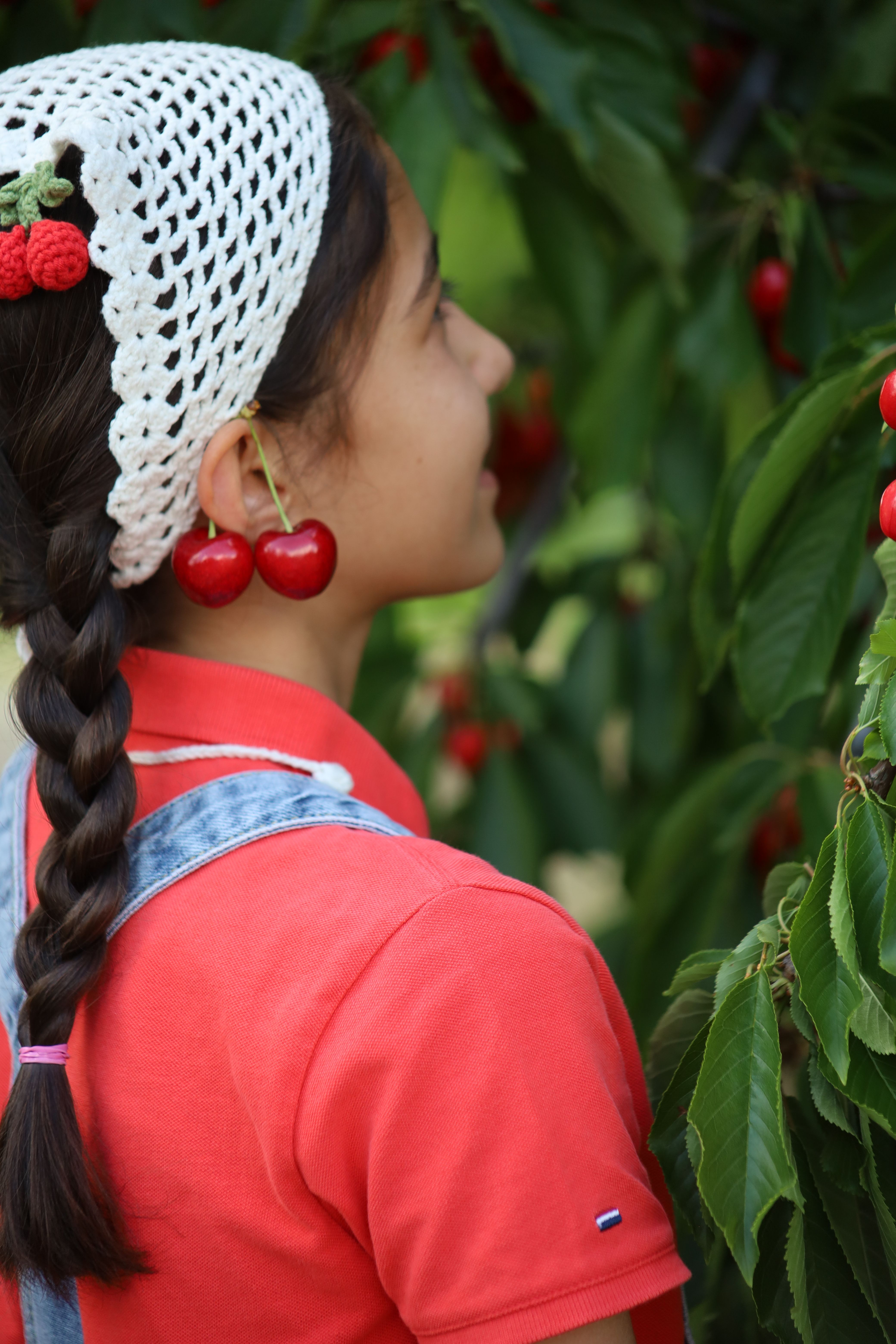 A girl with cherry earrings stands in a lush orchard.