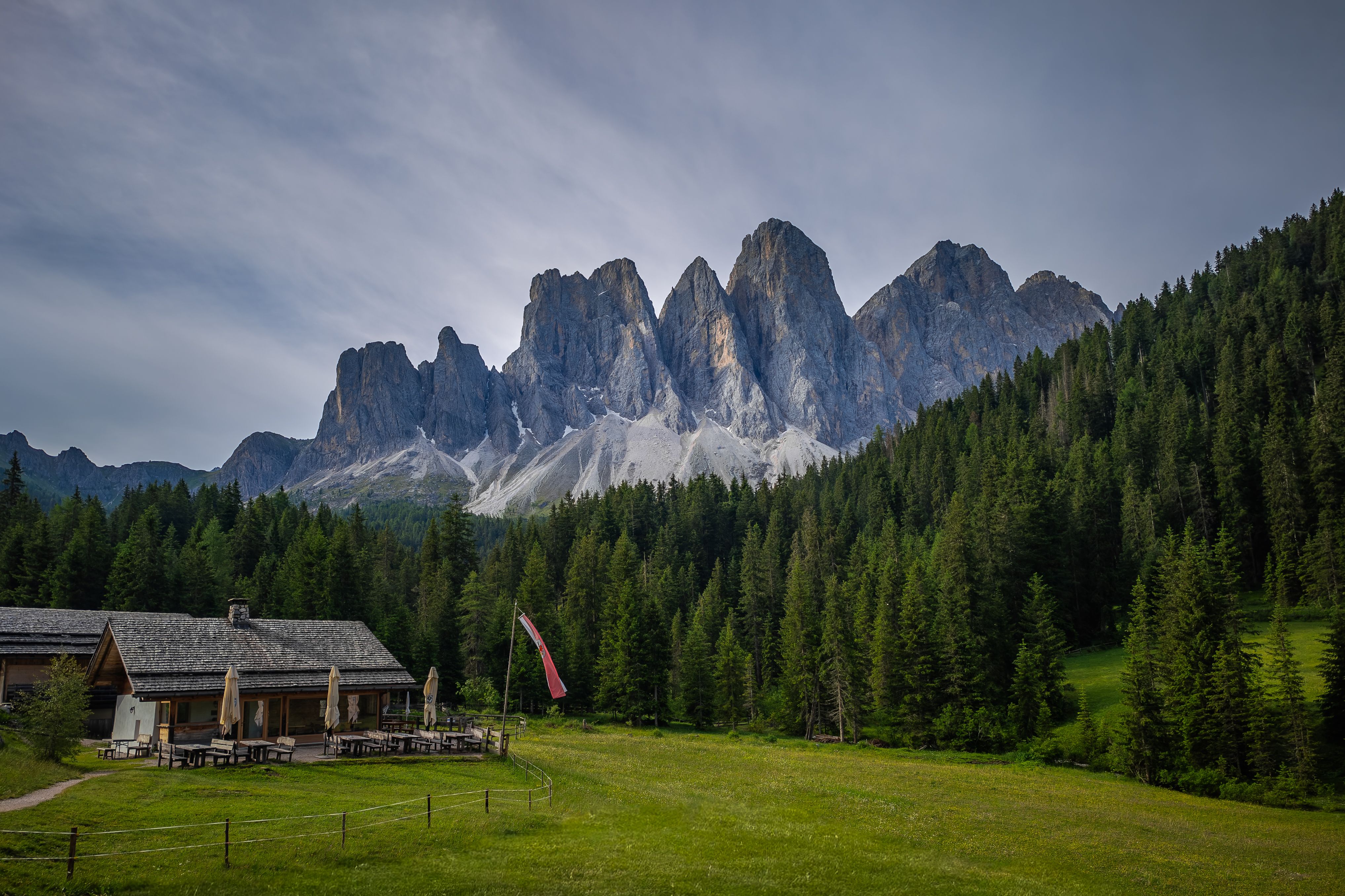 A serene view of a rustic cabin with the Dolomites in the background, surrounded by lush forest.