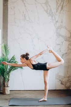 Elegant woman practicing a yoga pose indoors with a marble wall backdrop, showcasing flexibility and balance.