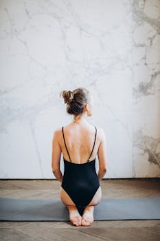 一名女子身穿黑色泳衣，在室内坐在瑜伽垫上练习