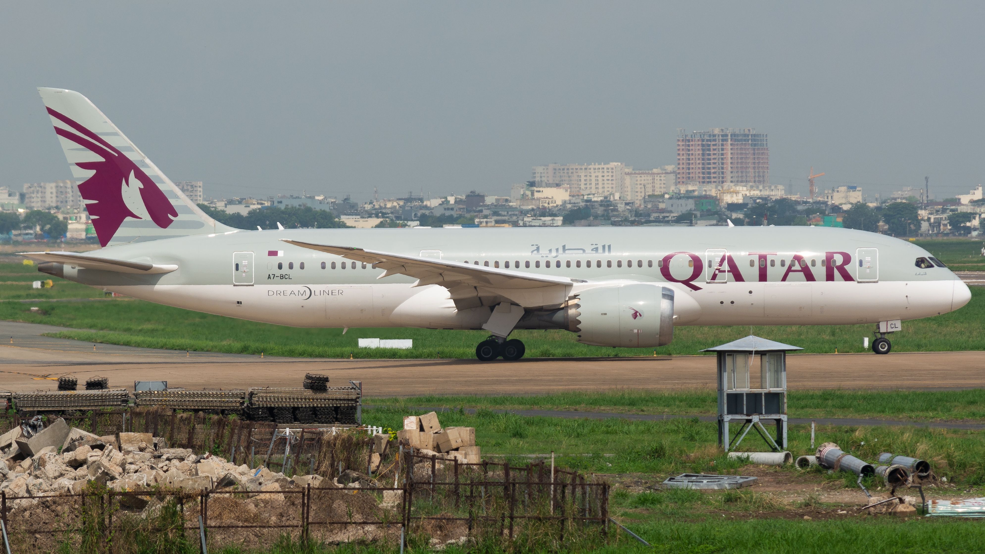 Ein Qatar Airways Dreamliner mit weinrotem Logo rollt auf der Startbahn eines Flughafens vor einer Stadtkulisse im Hintergrund.