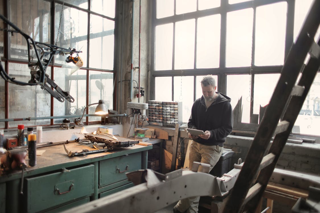 Adult bearded mechanic in casual wear standing near window in workshop and browsing digital tablet