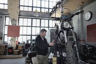 Side view of bearded adult mechanic in casual uniform standing near motorcycle and working with engine in workshop