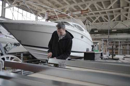 Concentrated bearded adult workman in casual wear working with metal detail near boat in garage