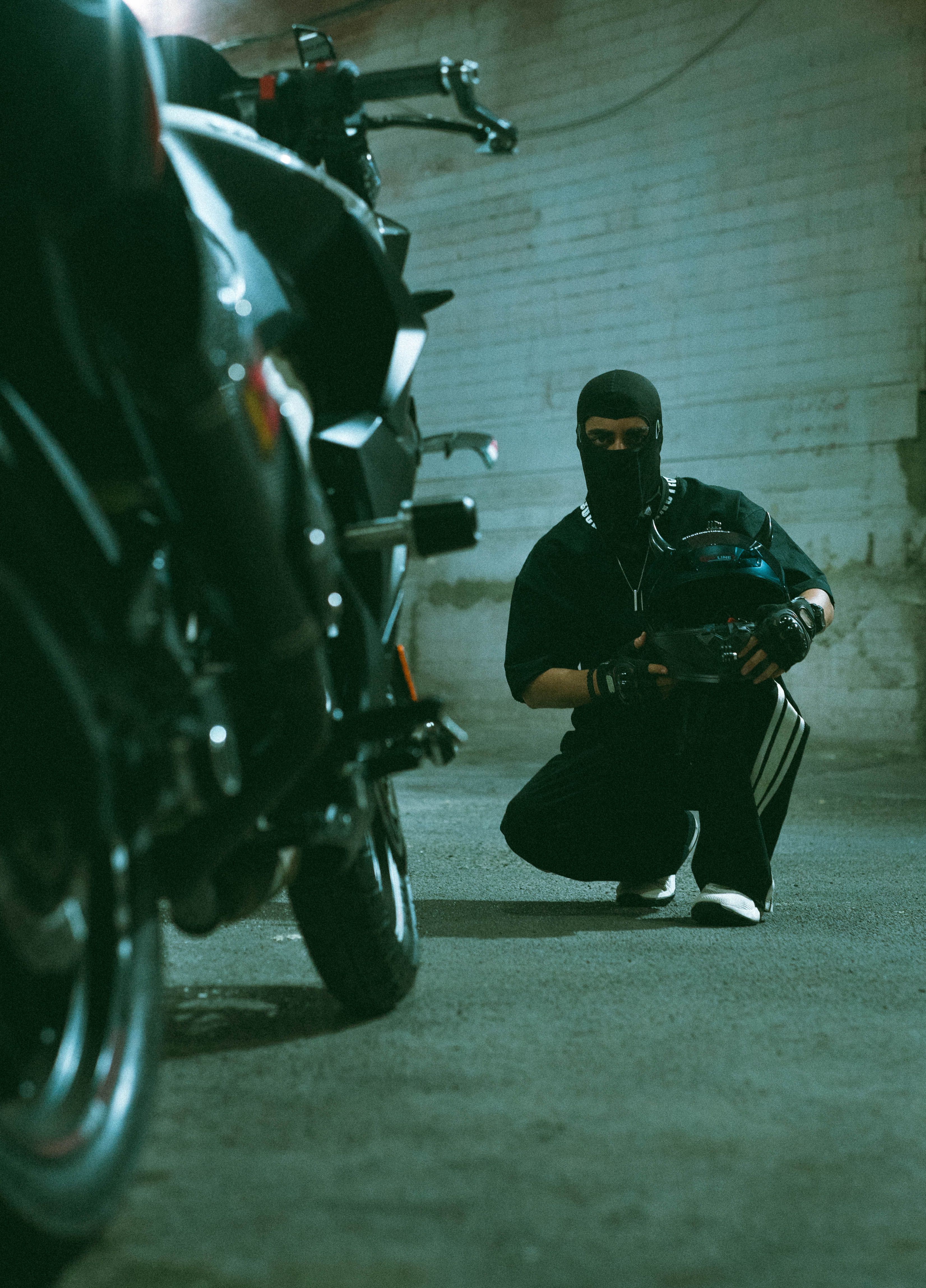 A masked person in a dimly lit garage crouching near a motorcycle, creating a mysterious scene.