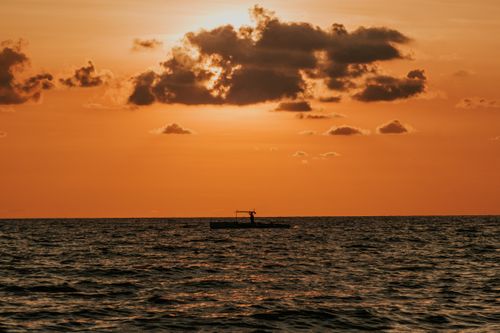 Free Silhouette of a boat against a vibrant orange sky reflecting on the sea at sunset, offering a serene scene. Stock Photo