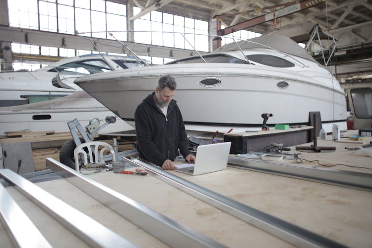 Adult Worker Using Laptop At Workbench During Work In Boat Garage