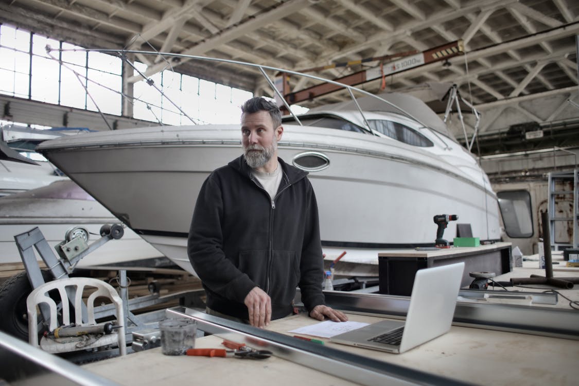 Adult bearded man in casual clothes using laptop while working with metal details near boat in workshop and looking away