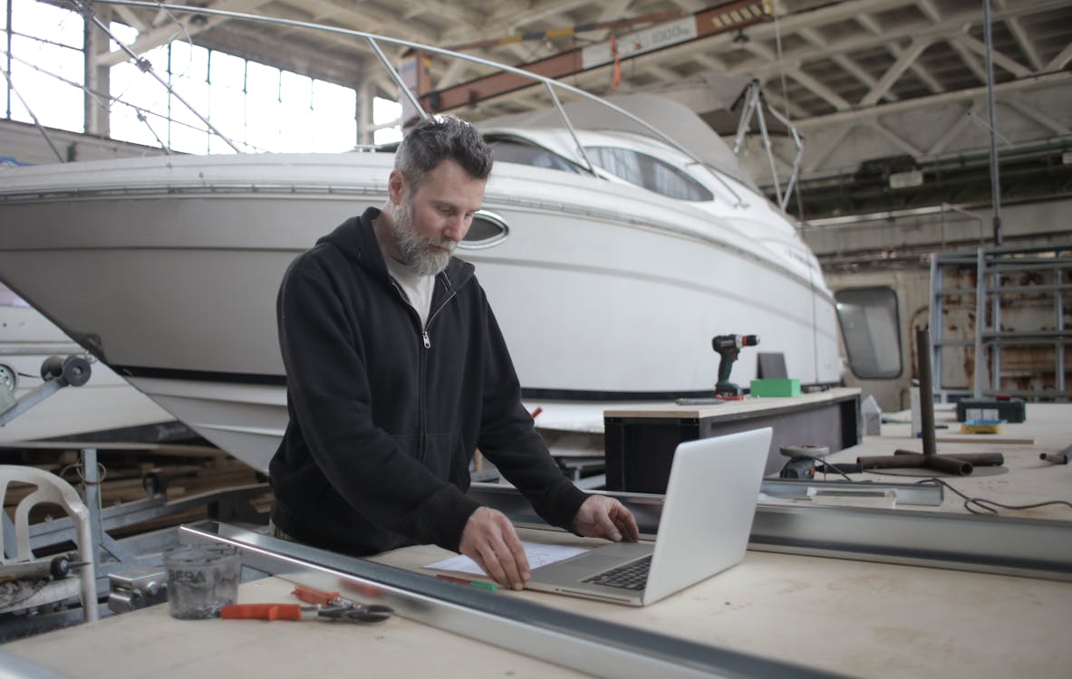 Nachdenklicher Erwachsener Arbeiter, Der Laptop Benutzt, Während Er Mit Metallteilen Nahe Boot In Der Werkstatt Arbeitet