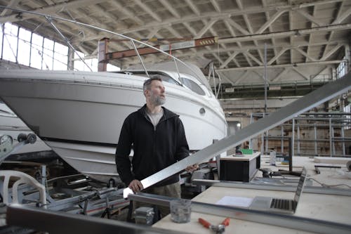 Side view of adult bearded workman in casual clothes standing near workbench and controlling metal detail in garage with yachts