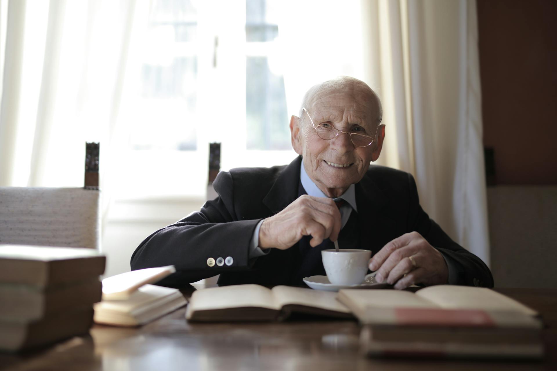 Un homme âgé en costume officiel buvant une boisson chaude tout en étant assis à table avec un livre