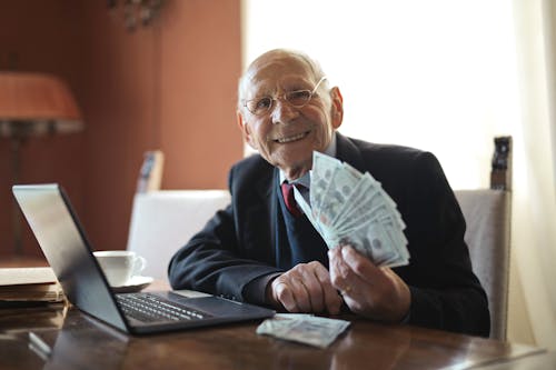 Heureux Homme D'affaires Senior Tenant De L'argent Dans La Main Tout En Travaillant Sur Un Ordinateur Portable à Table