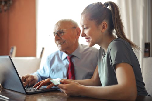 Free Young woman in casual clothes helping senior man in formal shirt with paying credit card in Internet using laptop while sitting at table Stock Photo