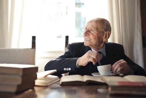 Positive senior man in formal suit and eyeglasses drinking hot beverage from cup while sitting at wooden table with books and looking away