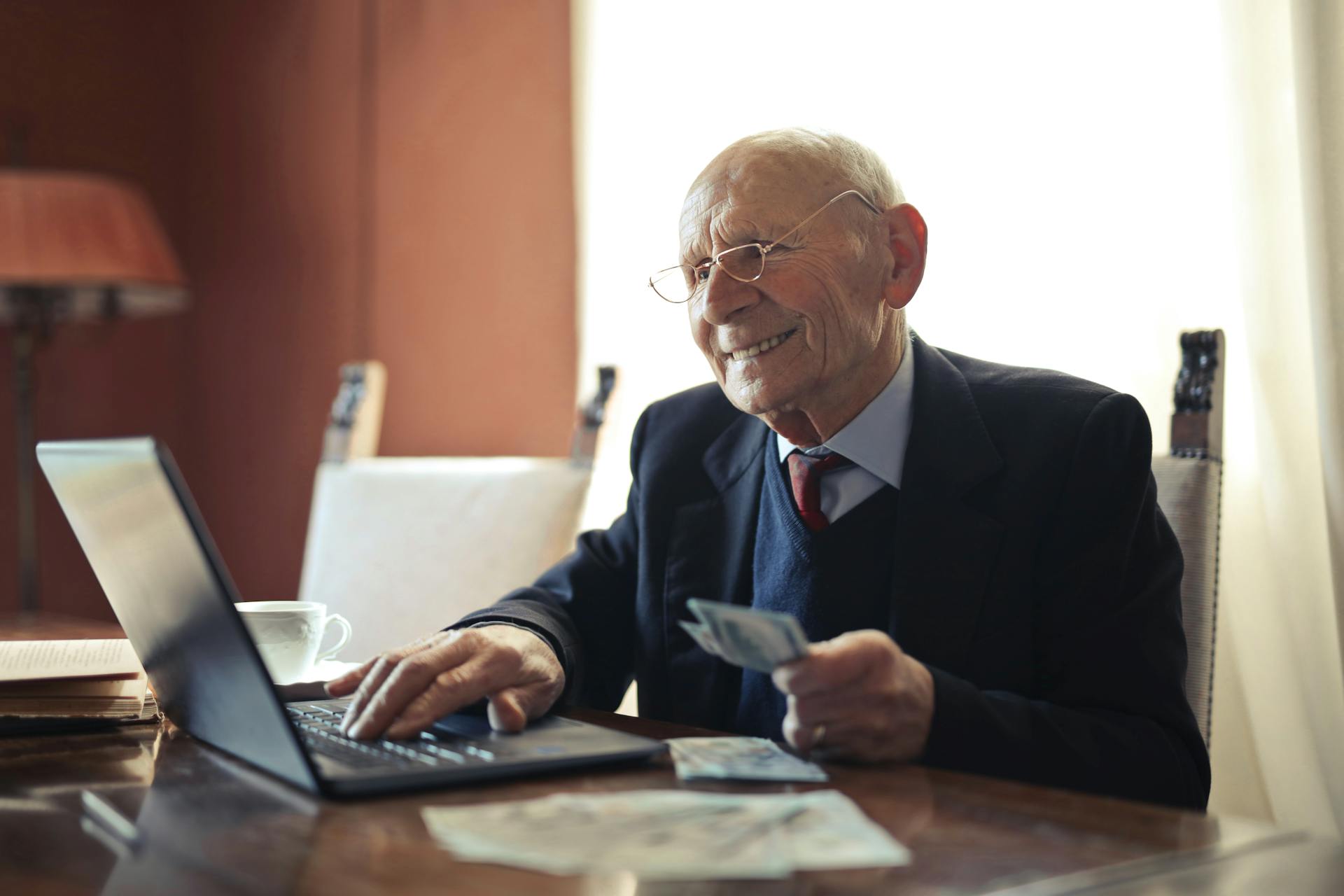 Positive senior businessman typing on laptop while holding money in hand