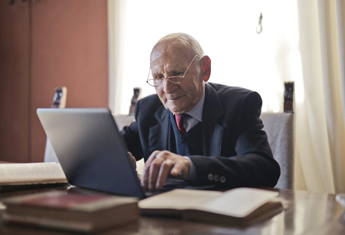 Poważny Starszy Mężczyzna Za Pomocą Laptopa Siedząc Przy Stole Z Książkami